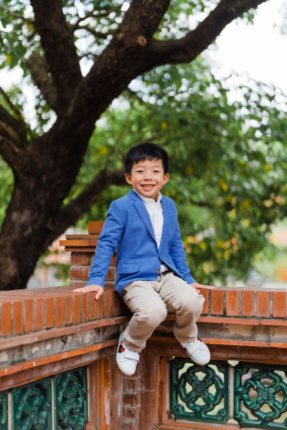 Child in bright blue blazer sitting cheerfully on brick wall under large tree.