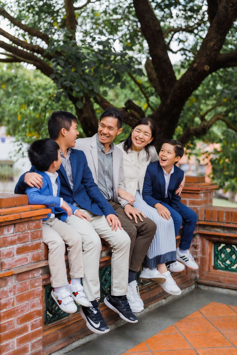 Family of four sharing laughter while sitting together on a brick wall under trees.