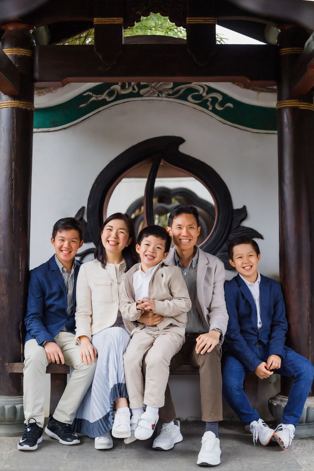 Happy family of five sitting together on wooden bench under ornate Asian architectural details.
