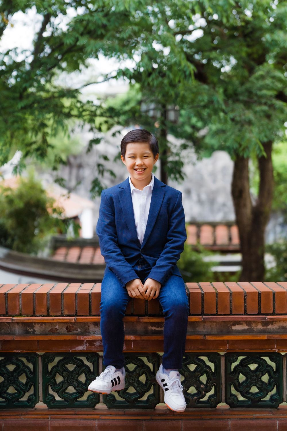 Young person in blue suit sitting on ornate brick wall in garden setting.