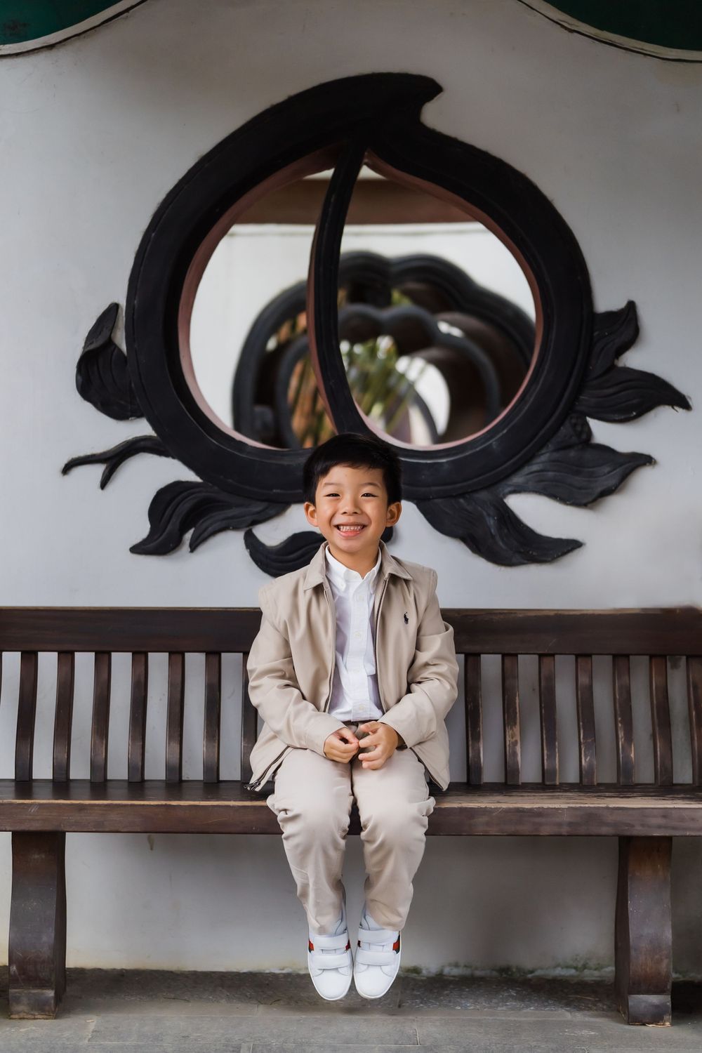 Child in beige suit sitting alone on bench under decorative circular wall design.