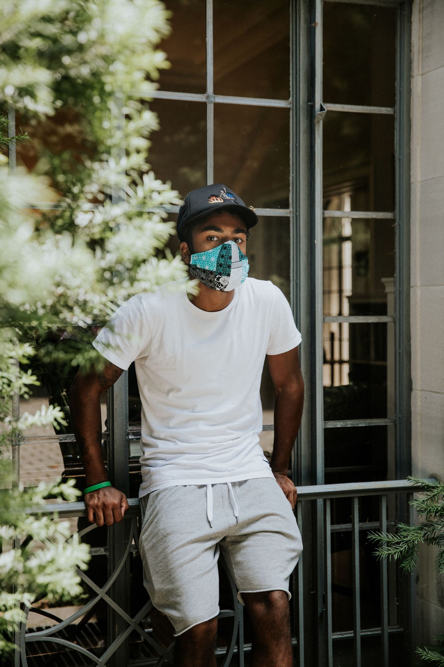 Person wearing blue face mask and white outfit sitting on porch with plants.
