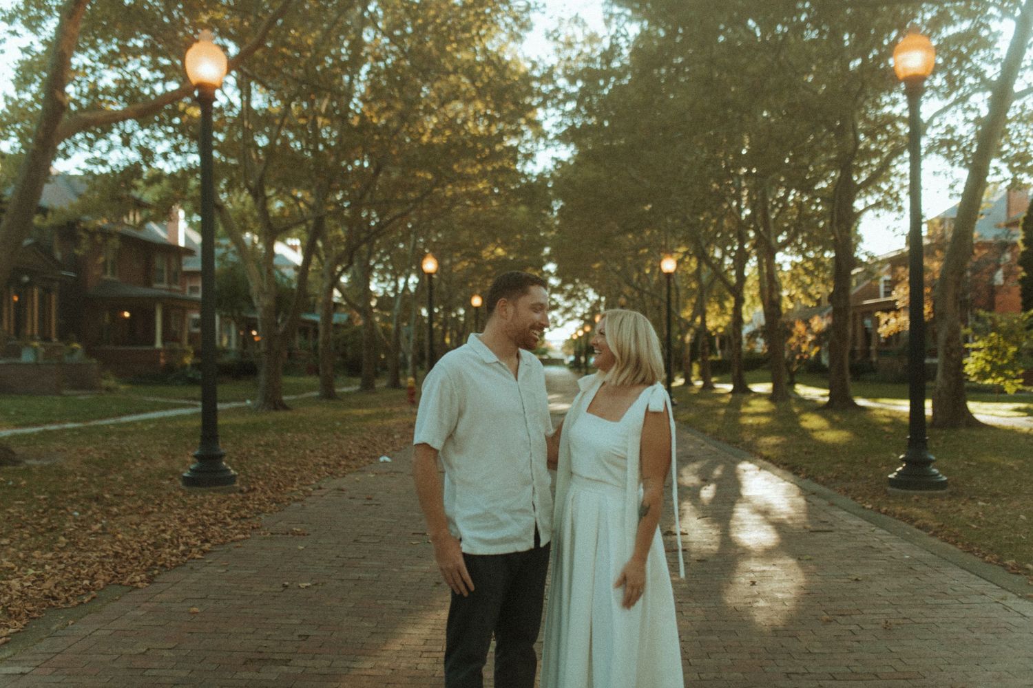 A romantic couple in white outfits shares a tender moment on a tree-lined path at dusk.