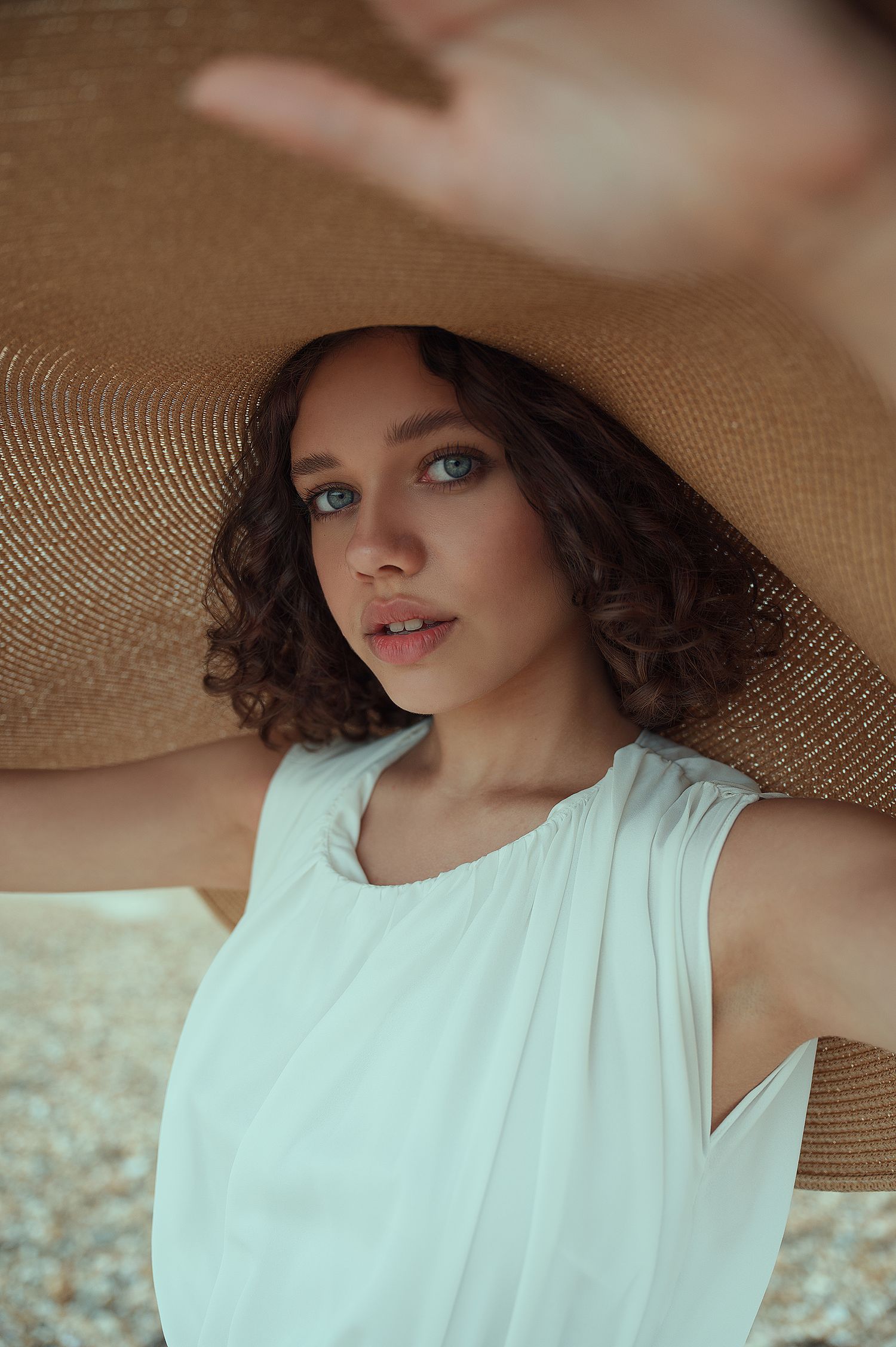 Portrait under wide brim hat shows soft makeup and natural curls against warm background.