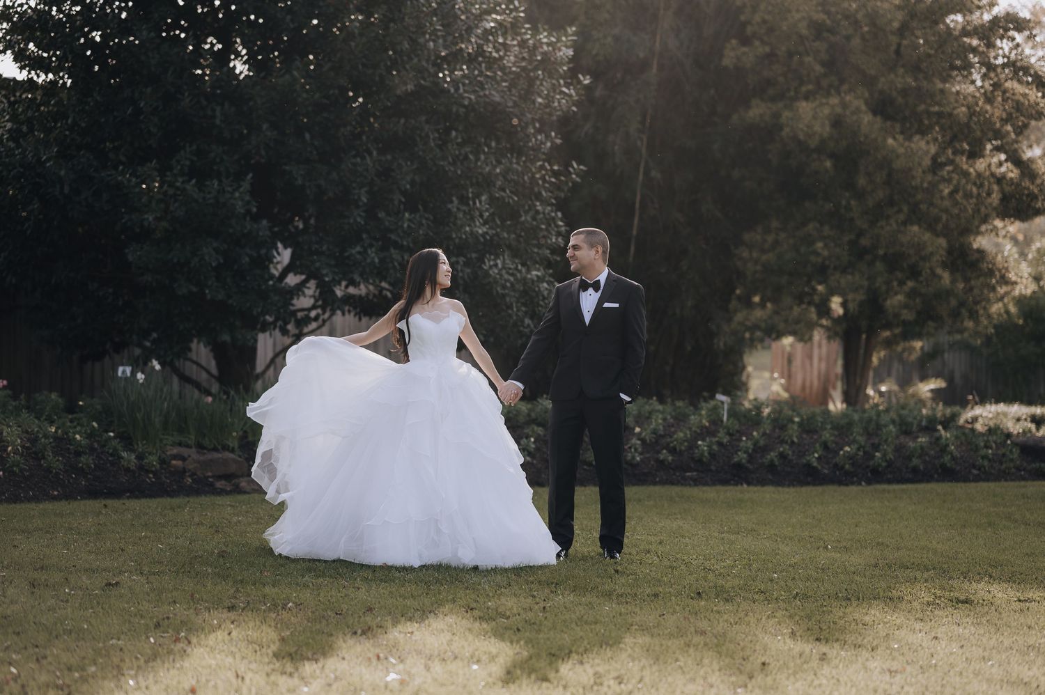 Newlyweds walking together on lawn with dramatic backlighting from setting sun.