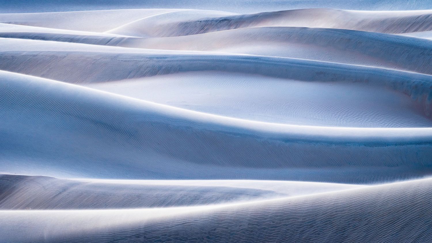 Abstract patterns of snow drifts creating smooth blue-tinted curves and waves.