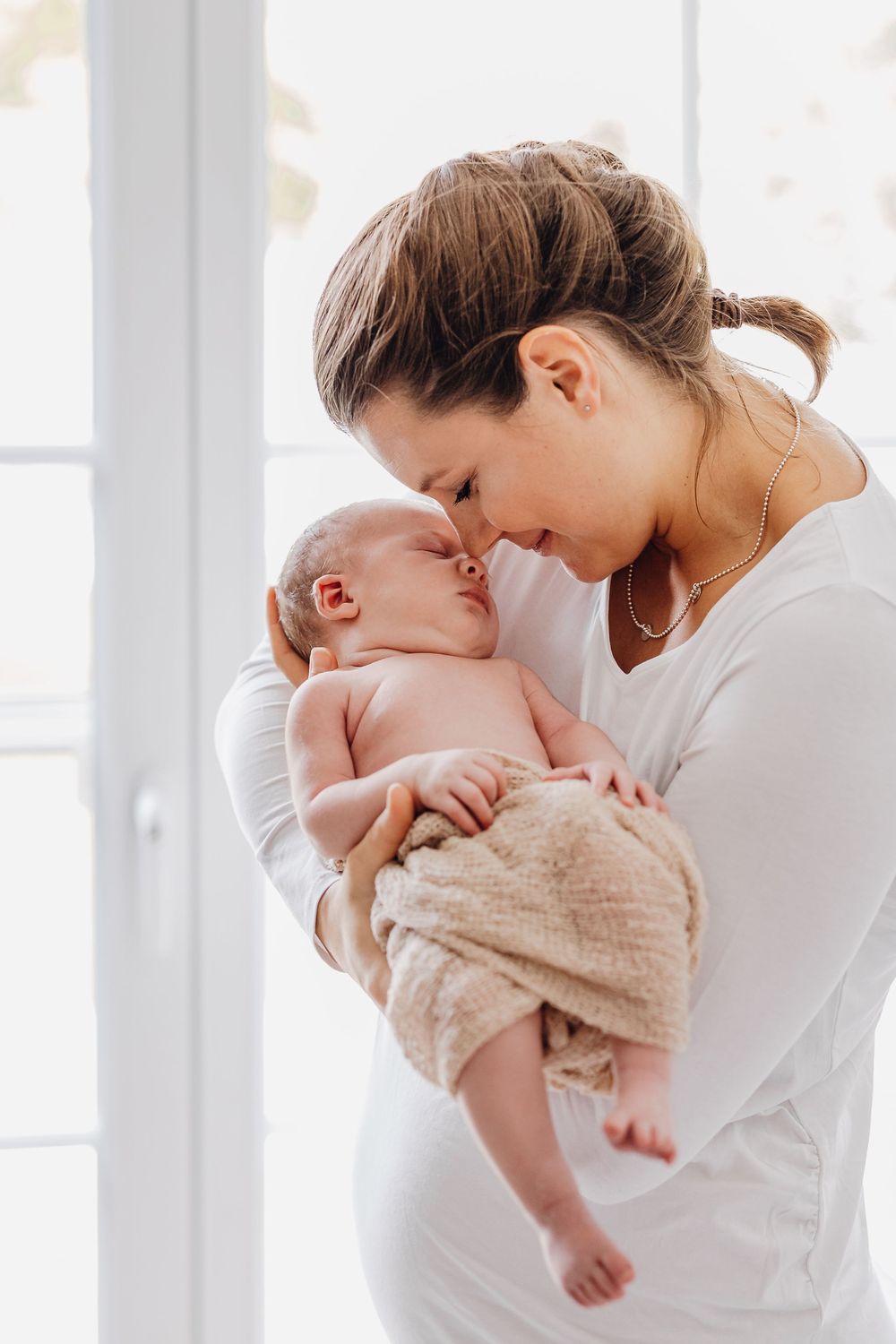 Elternteil küsst sanft neugeborenes Baby, während er am Fenster im weißen Raum steht.