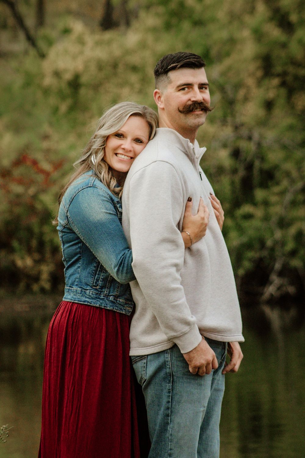A romantic couple poses by a lake, with her wearing denim and red while he sports a stylish mustache.