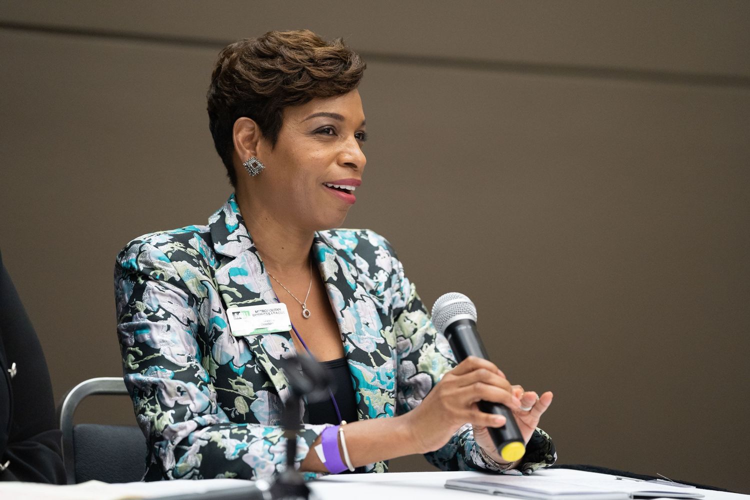 Confident woman in floral blazer speaks into mic at panel discussion, sharing insights during a professional conference or semina