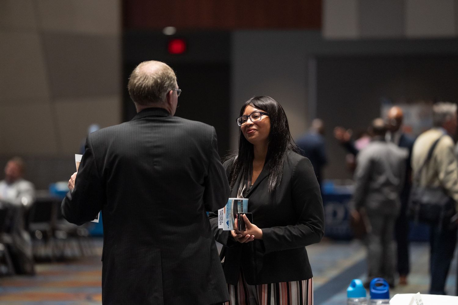 Professionals networking at a business conference, woman in blazer smiling and holding brochure during event conversation