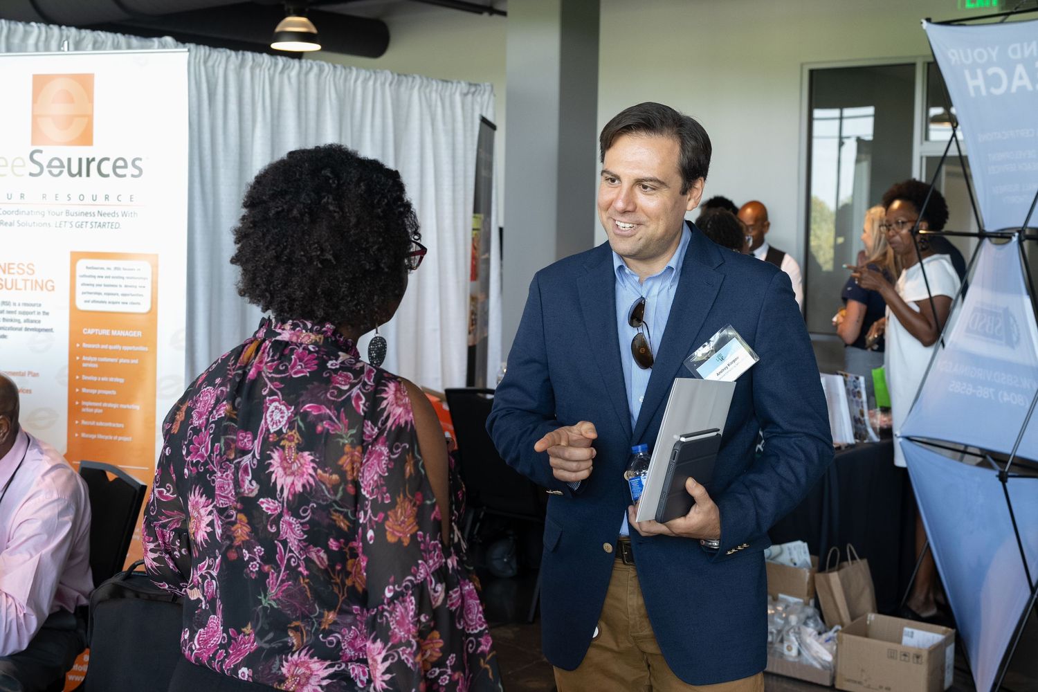 Man in navy blazer chats with woman at networking event, engaging in professional discussion near business resource booth