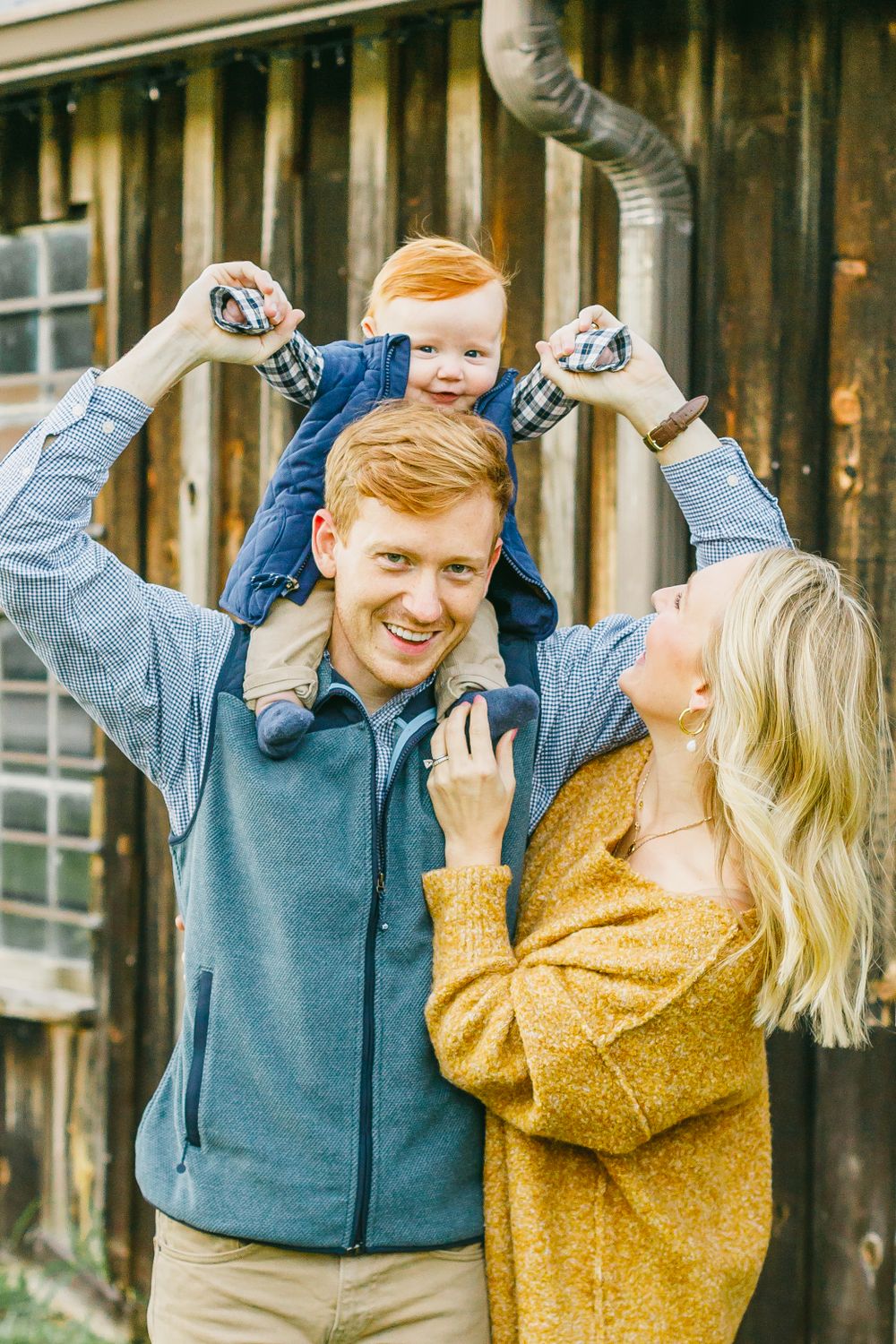 a man balances his redheaded baby on his shoulders, with mom looking up smiling