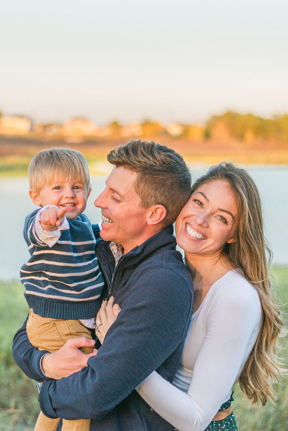 dad smiles at his two year old son in his arms, as his wife hugs him from behind