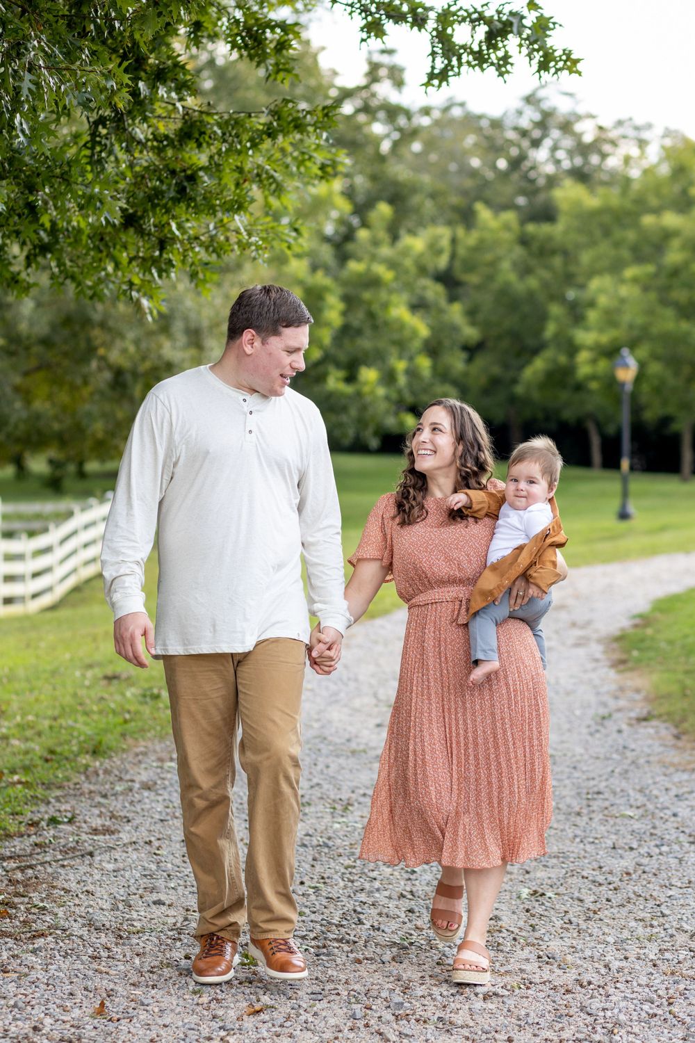 A couple in a pink dress and tan shirt walk in a green park holding hands with their infant son on mom's hip.