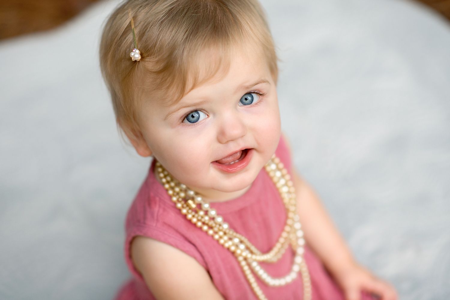 Smiling baby girl with blue eyes wearing a pink dress and pearl necklace with a flower hair clip.