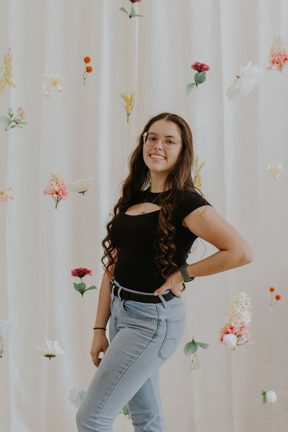 Portrait against floral backdrop of person in black top and jeans posing confidently.