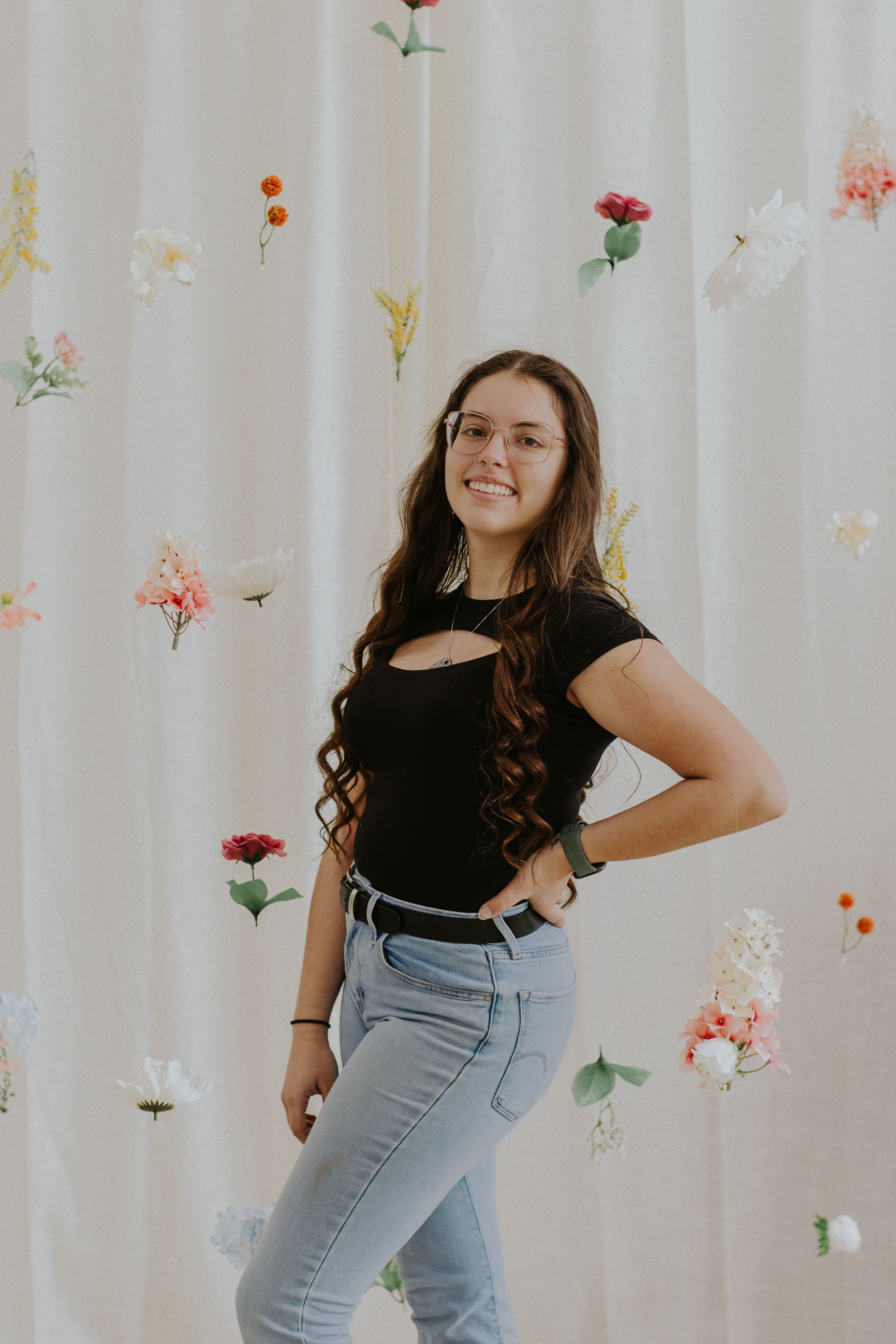 Portrait against floral backdrop of person in black top and jeans posing confidently.
