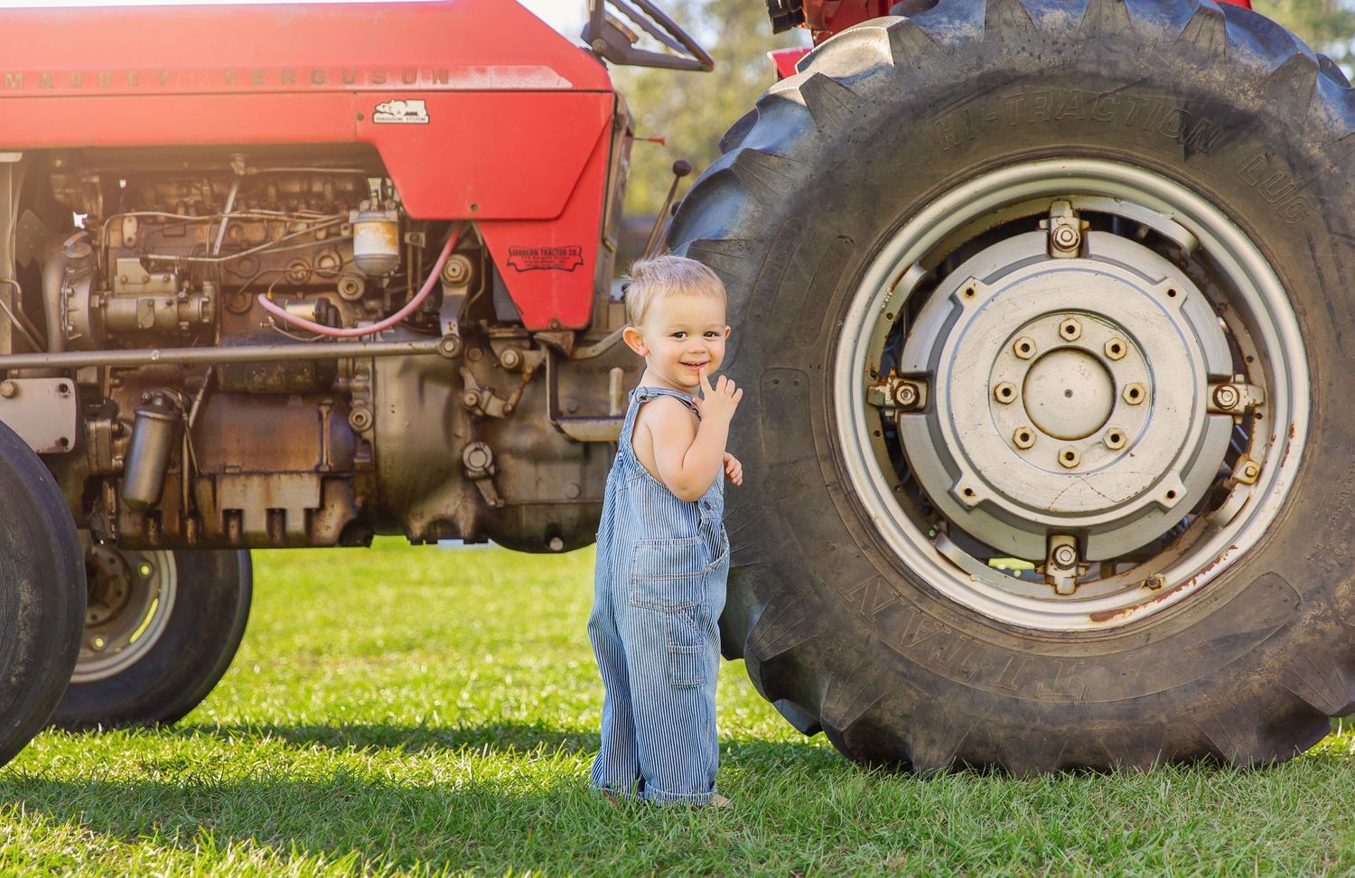 A small child in overalls standing next to a large red tractor wheel in a sunny field.