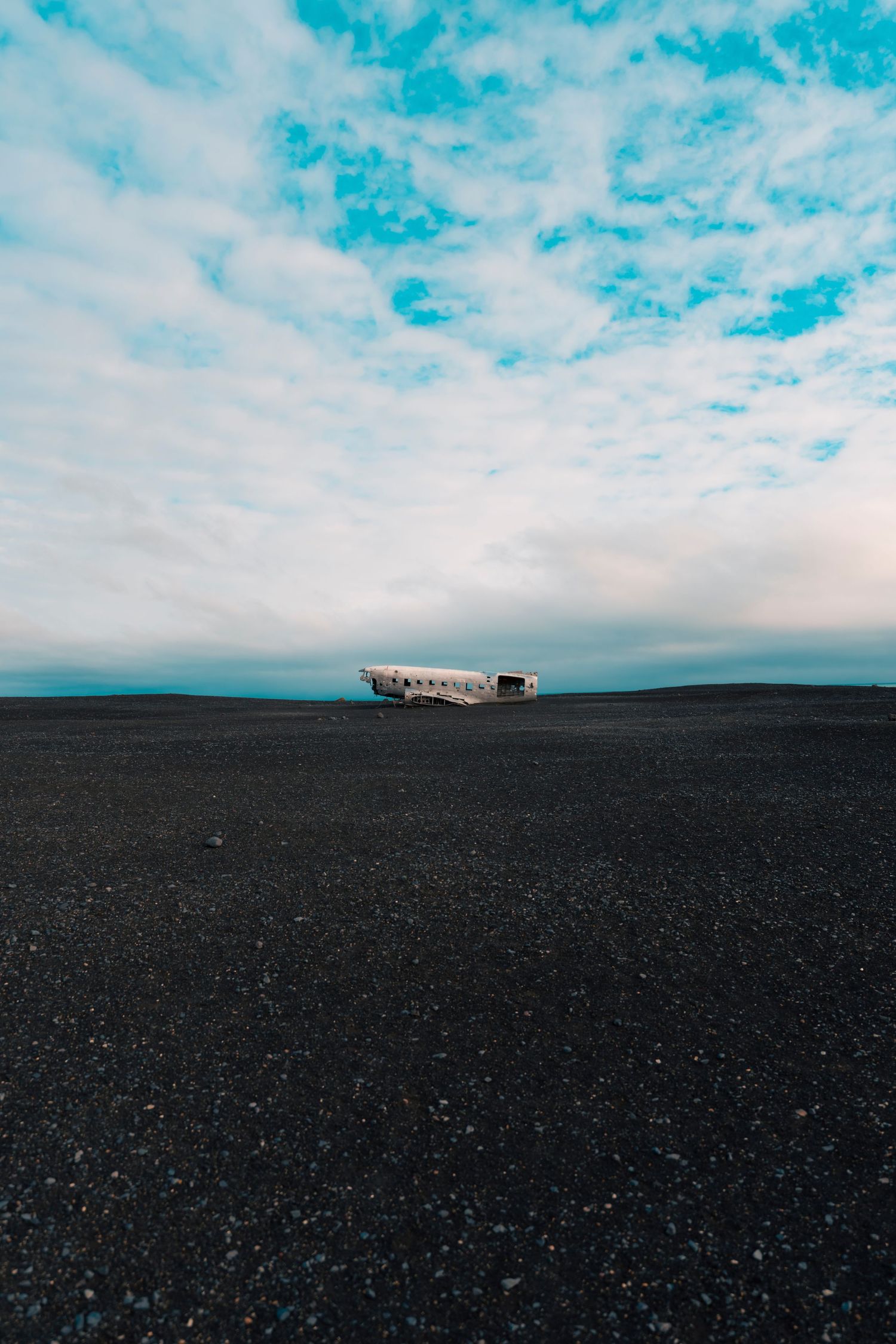 Abandoned plane wreckage on black volcanic sand under dramatic blue sky.