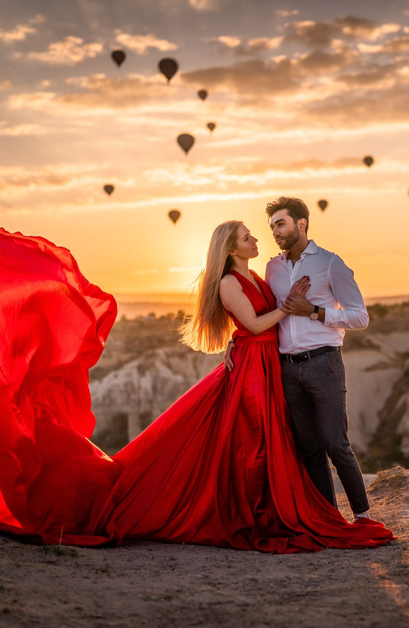 A flowing red dress catches the wind as a couple dances at sunset with hot air balloons overhead.
