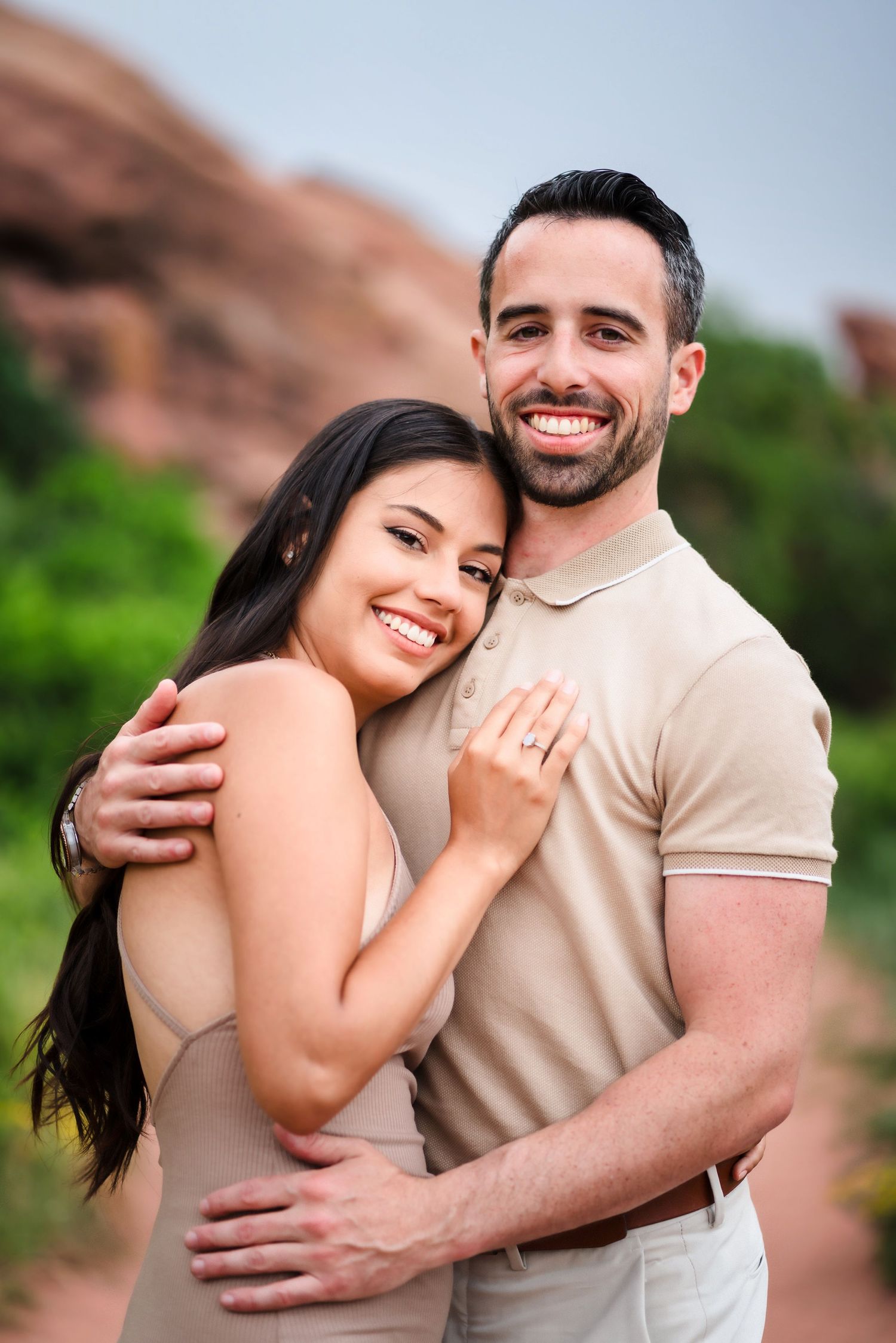 A couple in beige outfits embracing and smiling at Red Rocks Park with natural rock formations in the background.