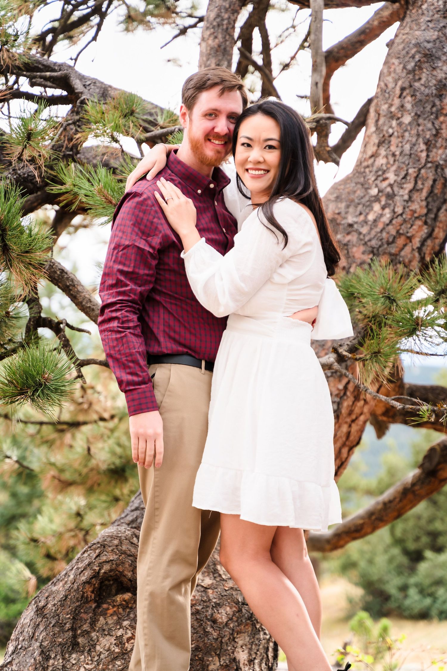 Couple posing together near a large pine tree with mountains visible in the distance.
