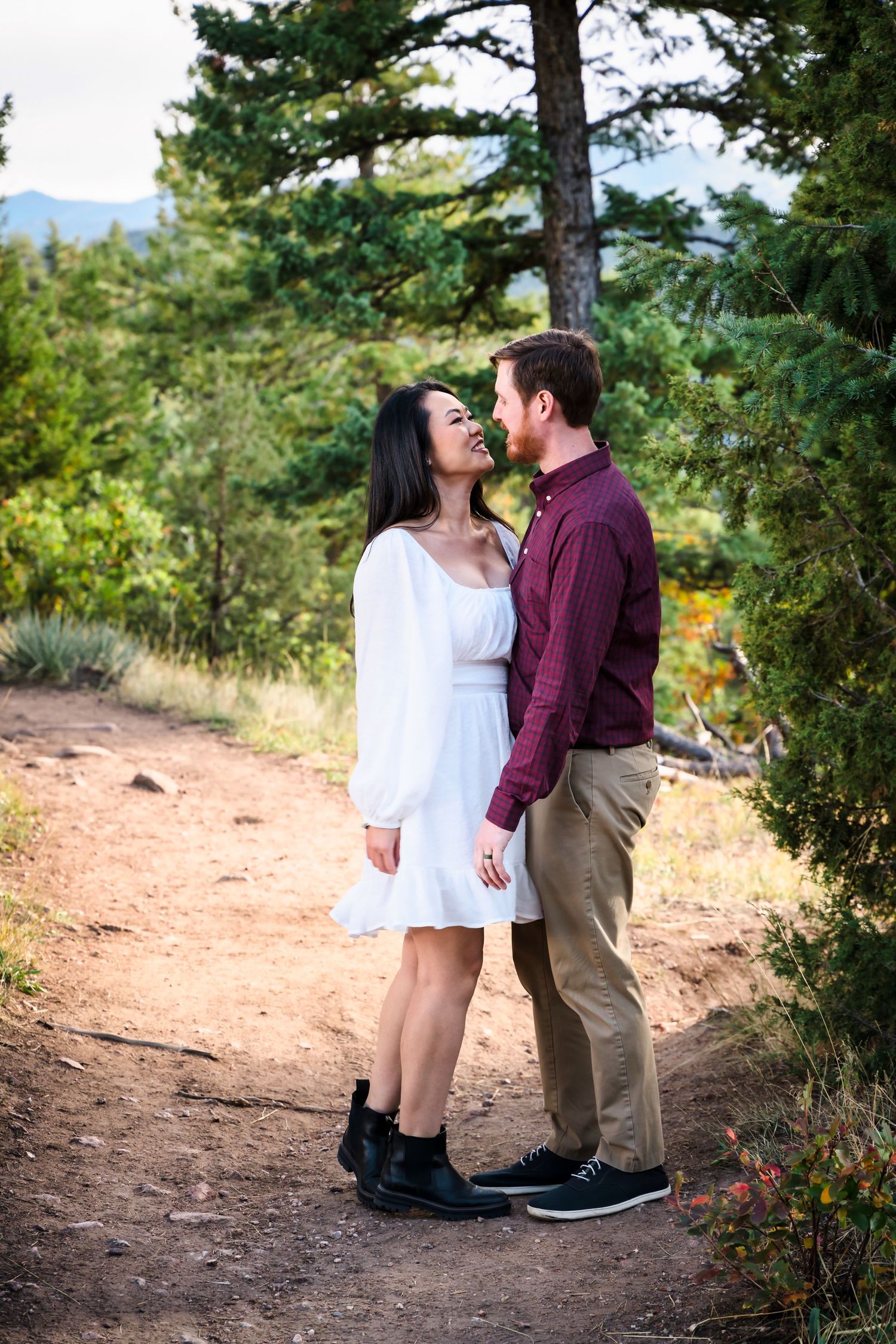 Couple standing close together on a dirt trail surrounded by pine trees.
