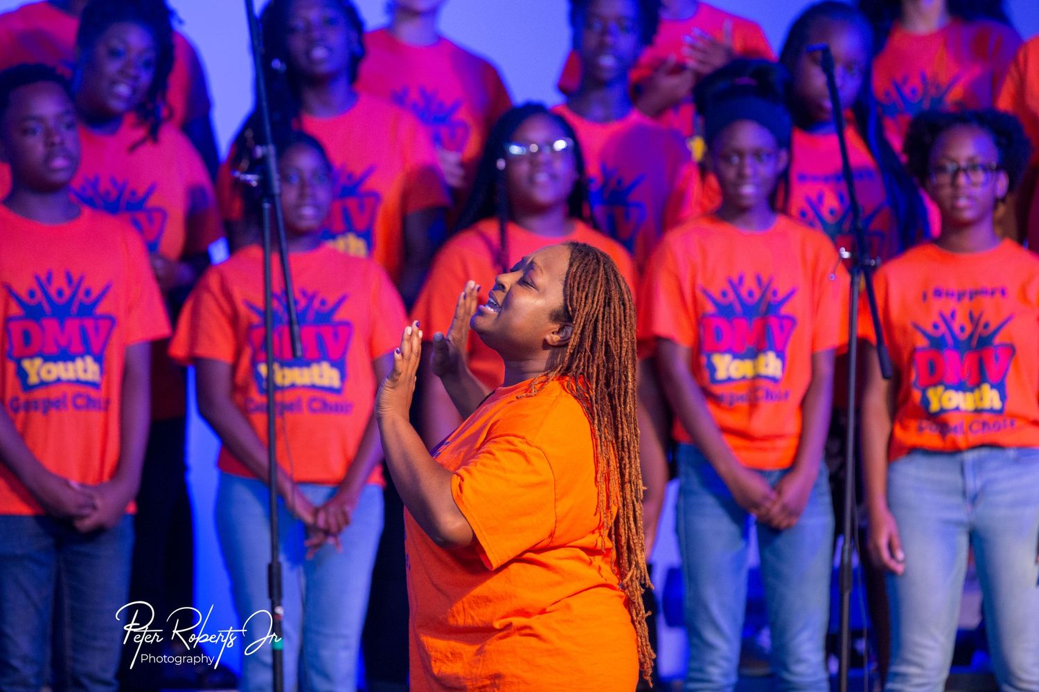 Passionate female soloist performing with youth gospel choir in matching orange shirts on stage.