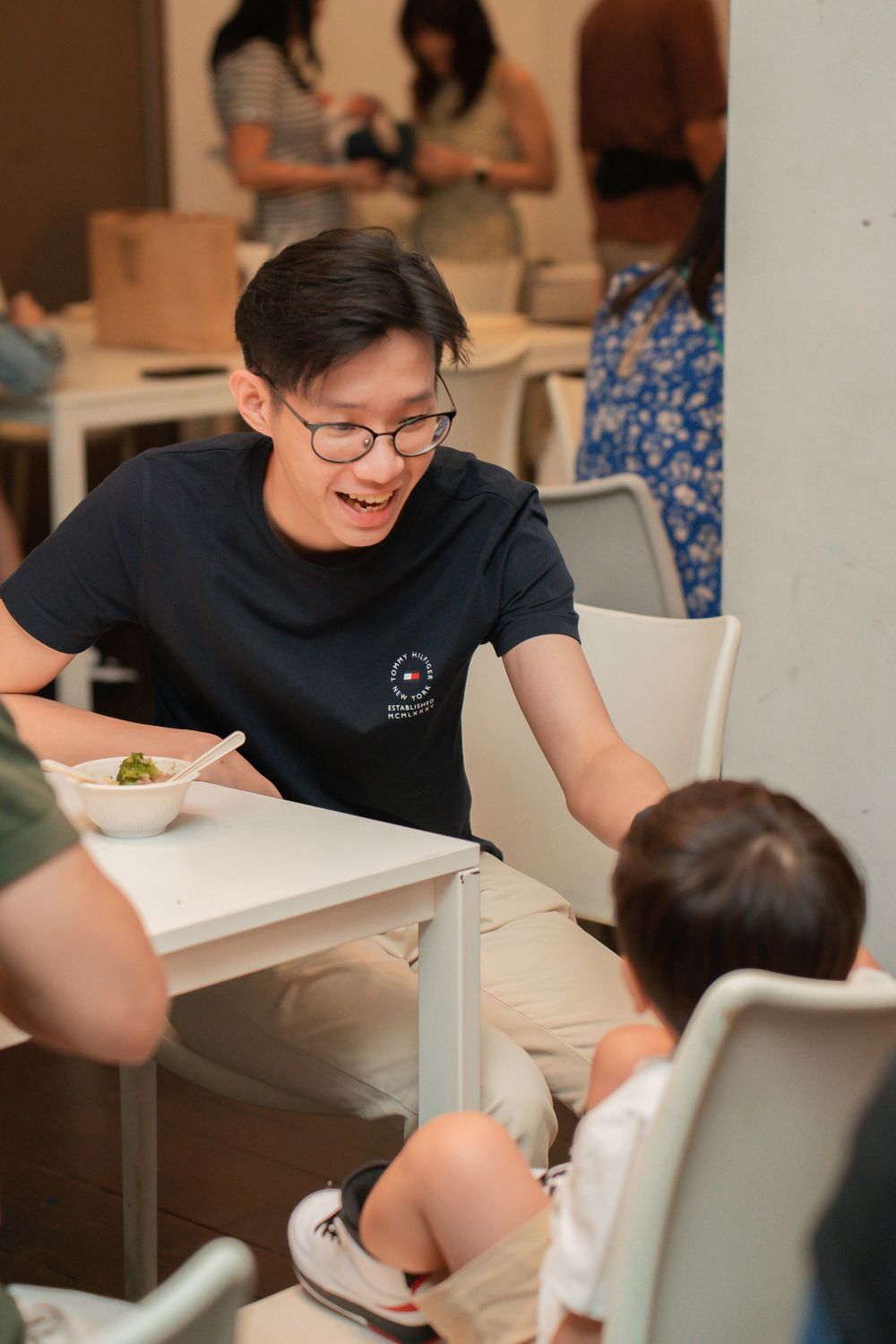 Person in glasses and black t-shirt laughs while interacting with a small child at a white table during a gathering.
