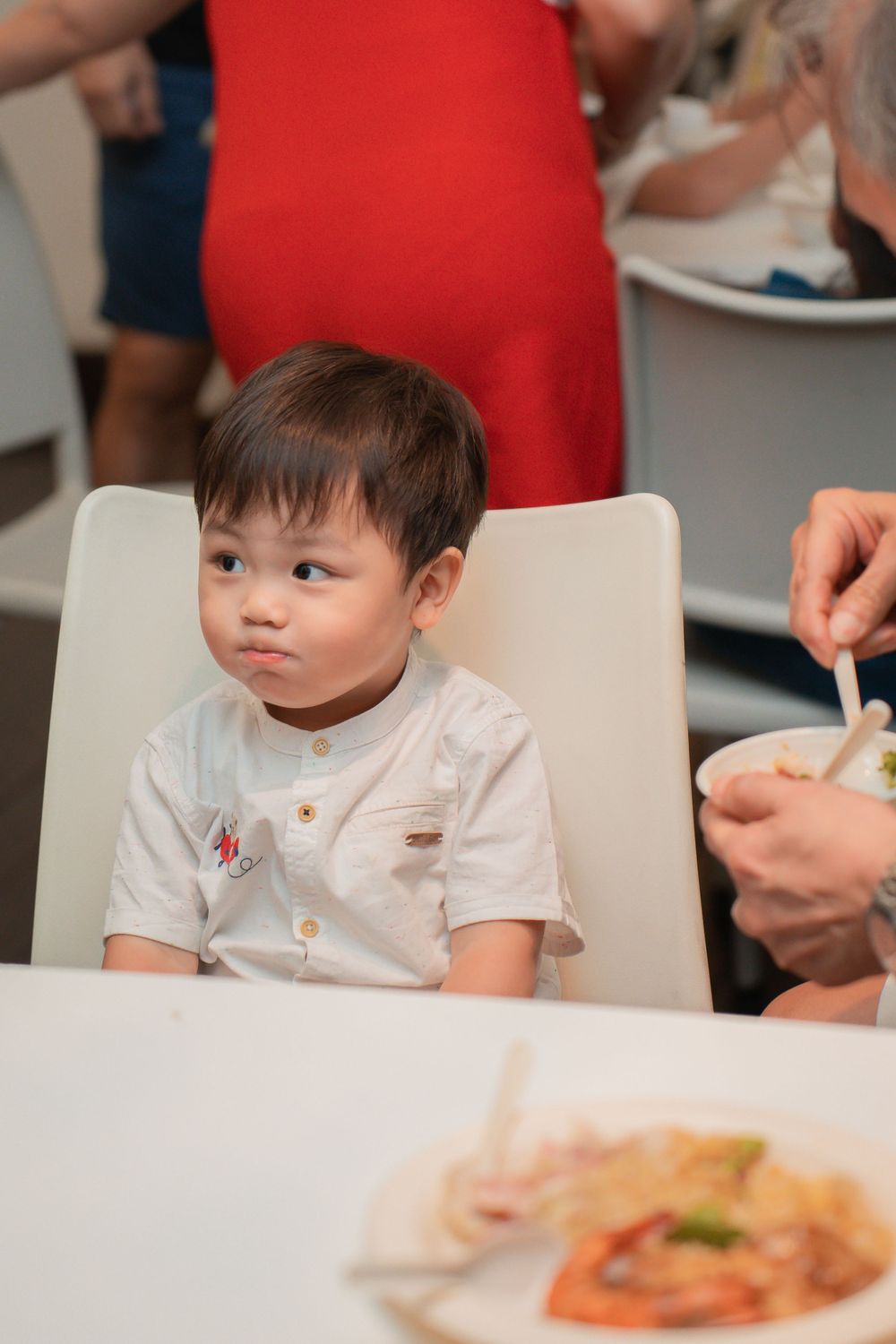 Small child in white shirt sits at table during meal time with curious expression.