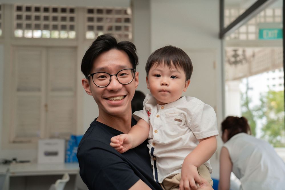 A young adult holding a toddler in a casual indoor setting with bright natural lighting.
