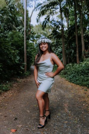 Graduate in silver dress poses playfully on forest trail.
