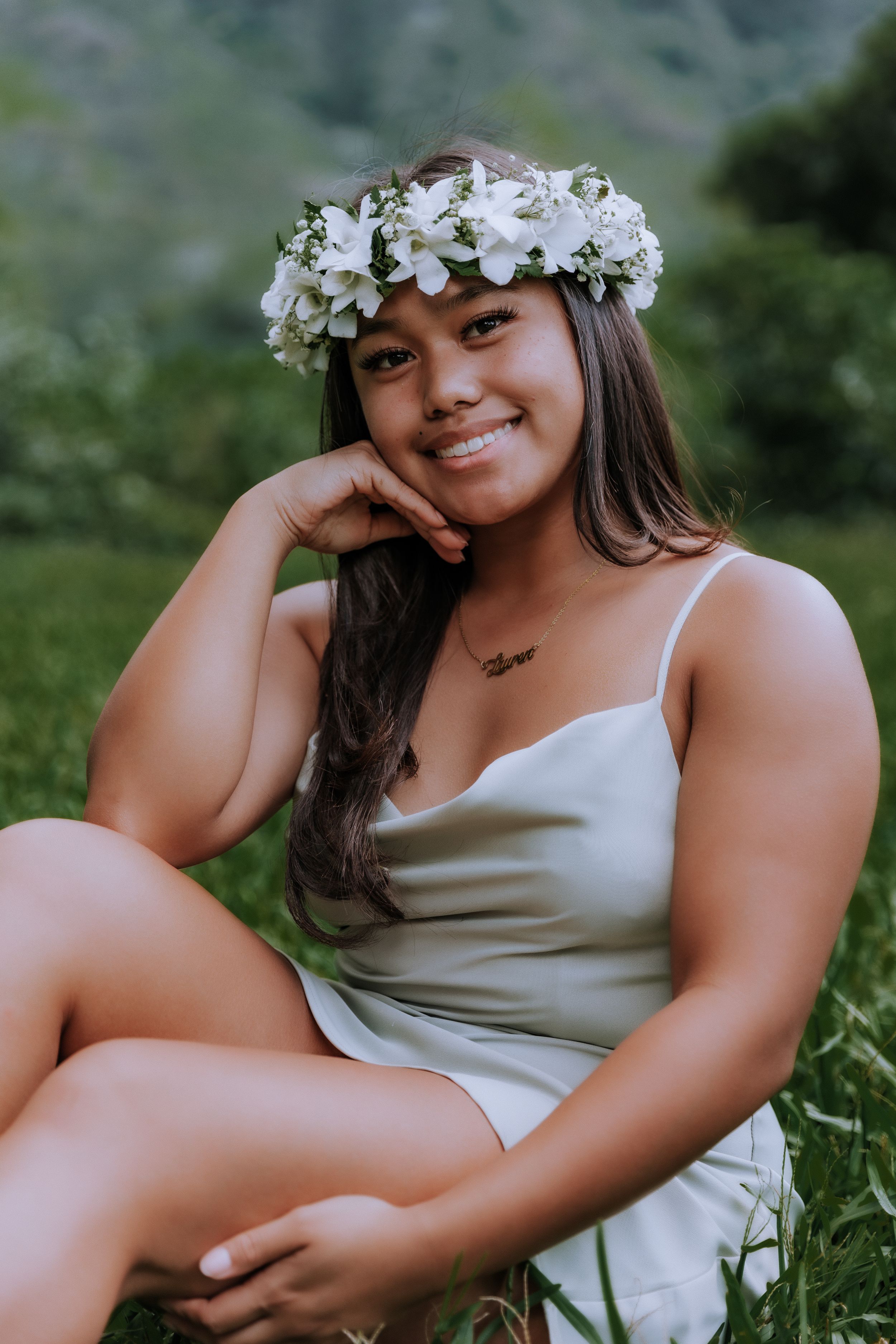 Graduate in silver dress and floral crown poses in grassy outdoor setting.