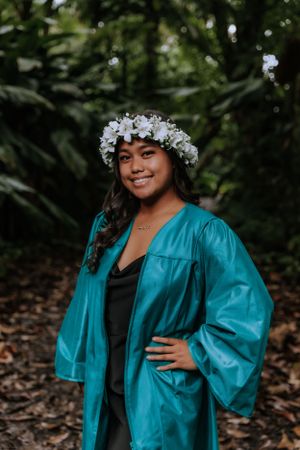 Graduate in teal gown and floral crown poses in outdoor setting.