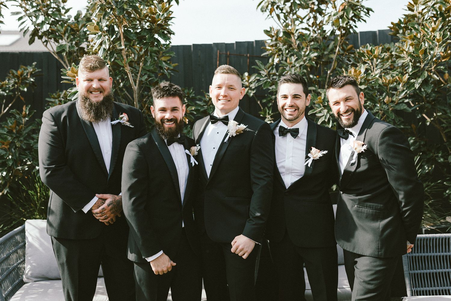 A group of men in formal black tuxedos pose together outdoors for a wedding photo.