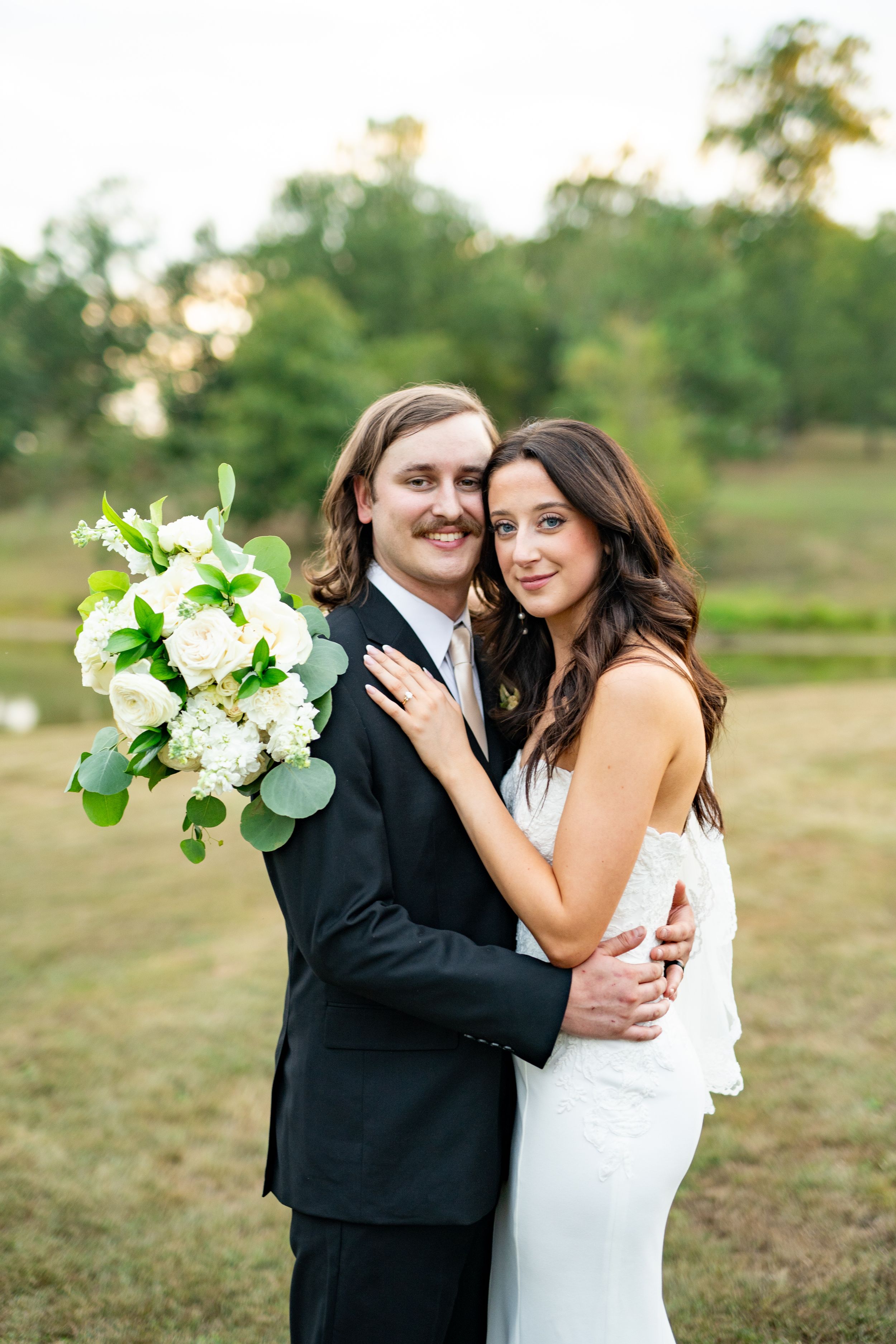 Two people in formal wedding attire embrace outdoors in natural setting with bouquet.