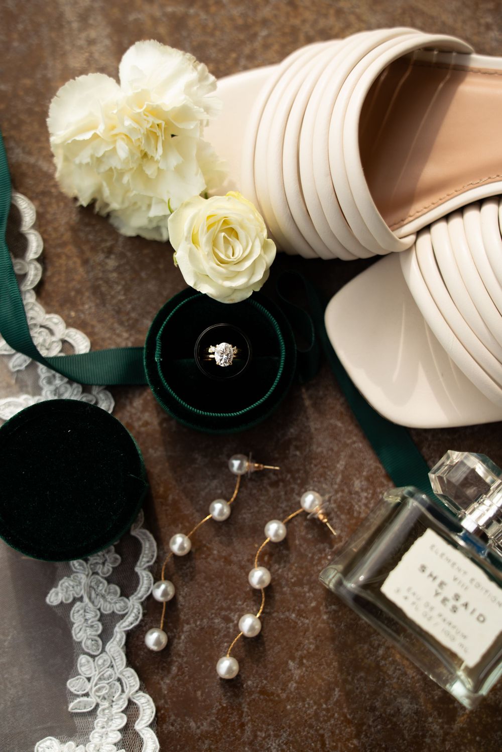 Wedding accessories including white shoes, roses, pearls and rings arranged on a dark rustic background.