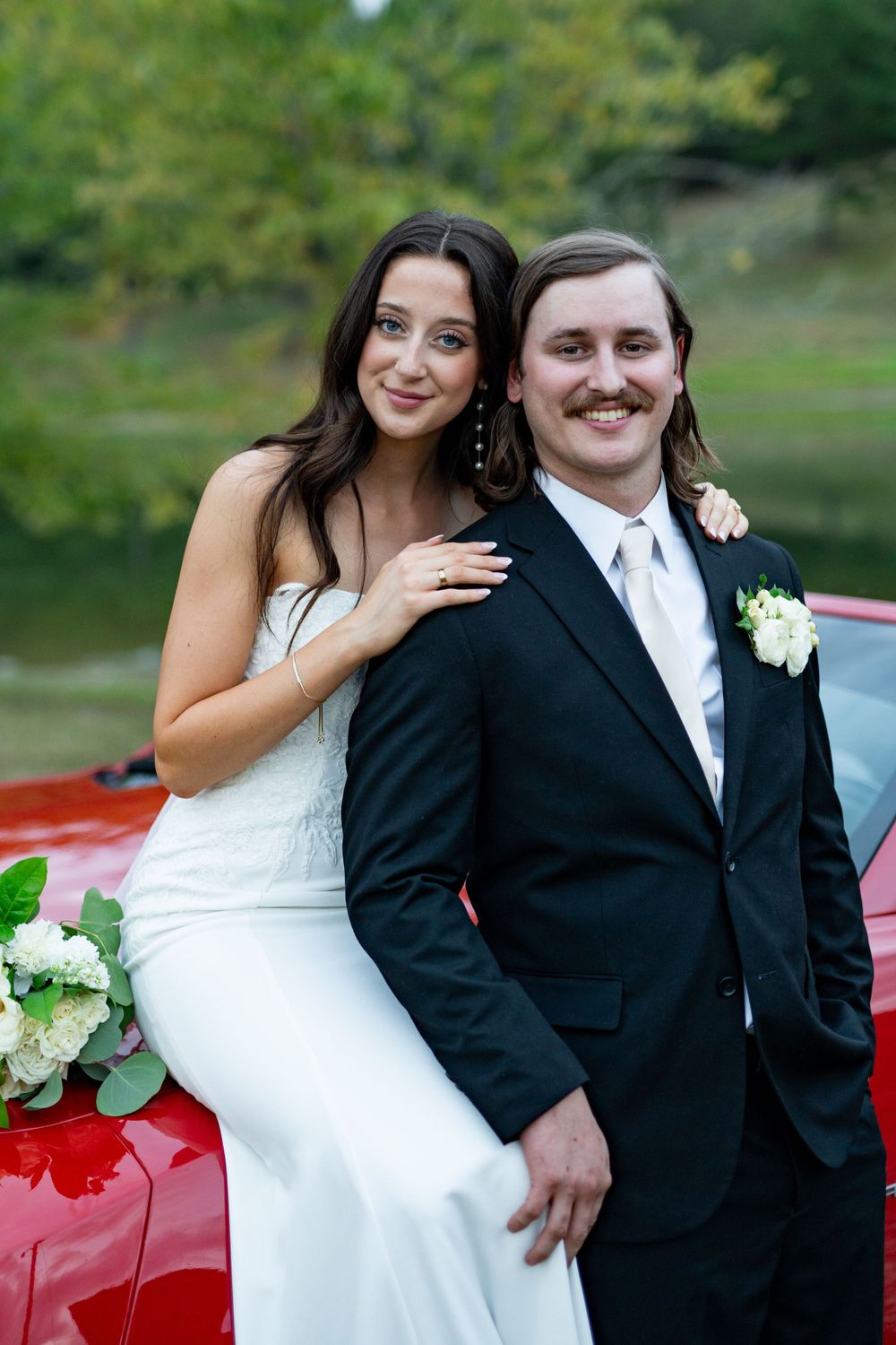 Wedding couple poses together against red car in outdoor setting.