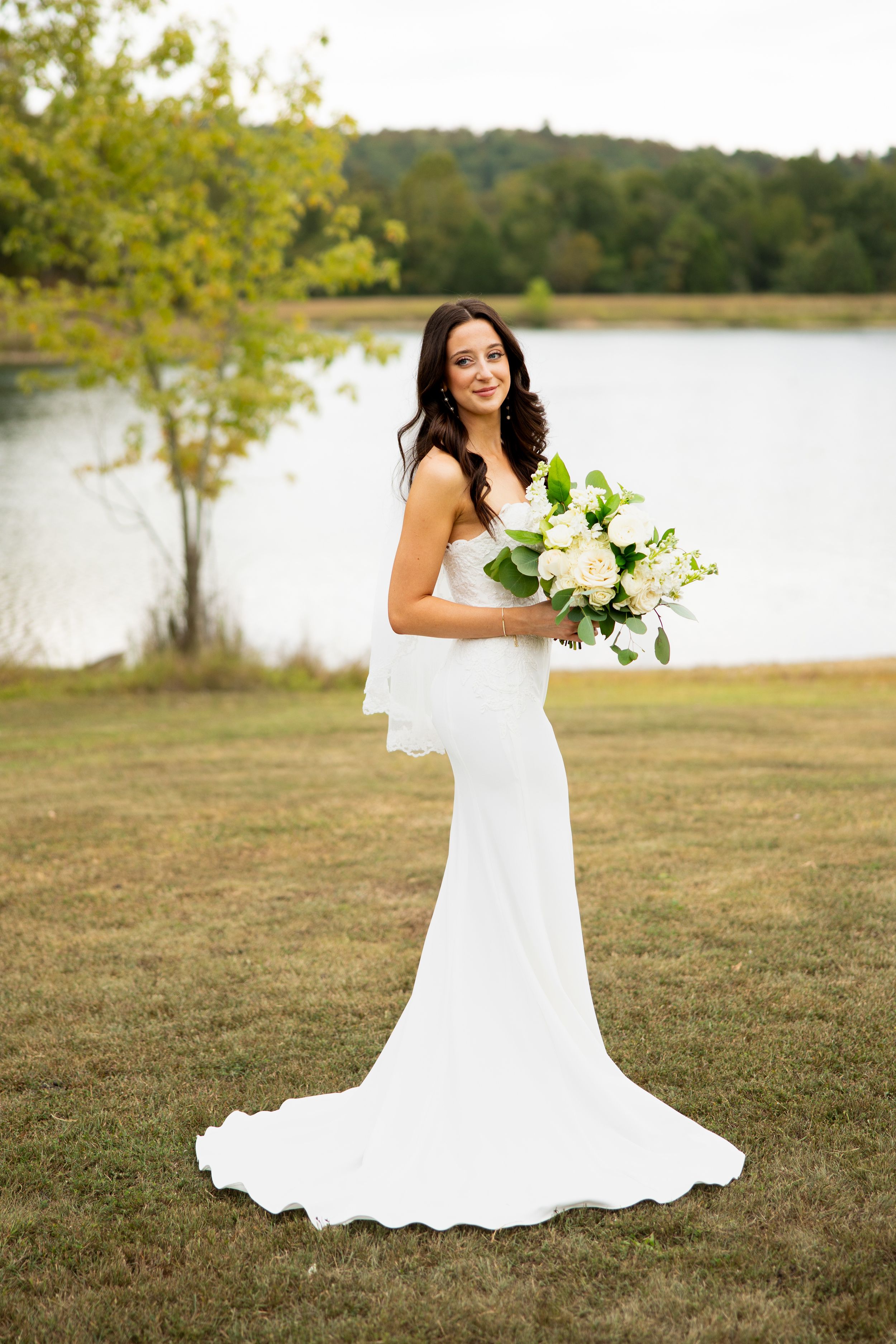 A bride in a white wedding gown poses with a bouquet against a lakeside backdrop.