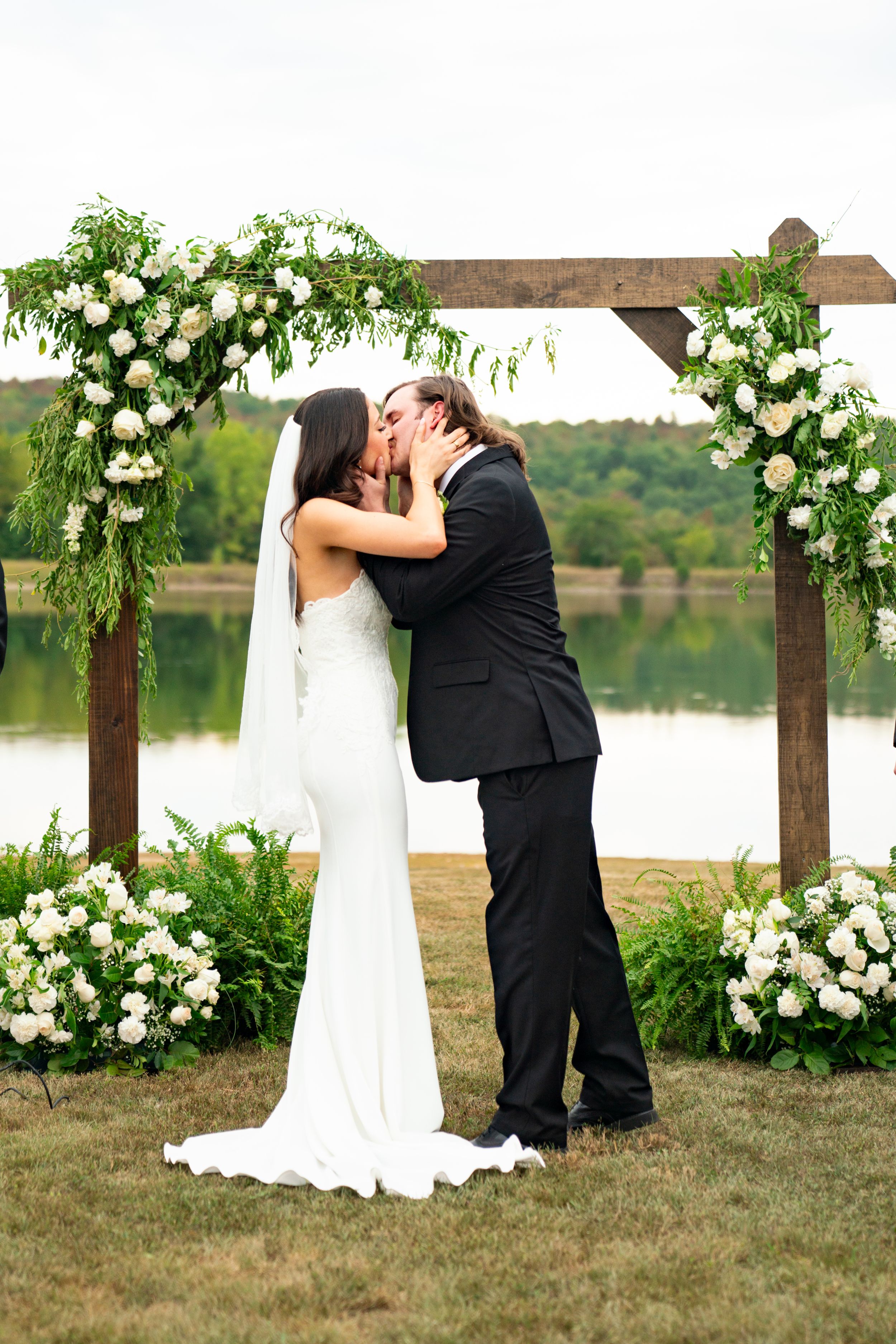 Newlyweds share their first kiss under a floral arch by the lake.