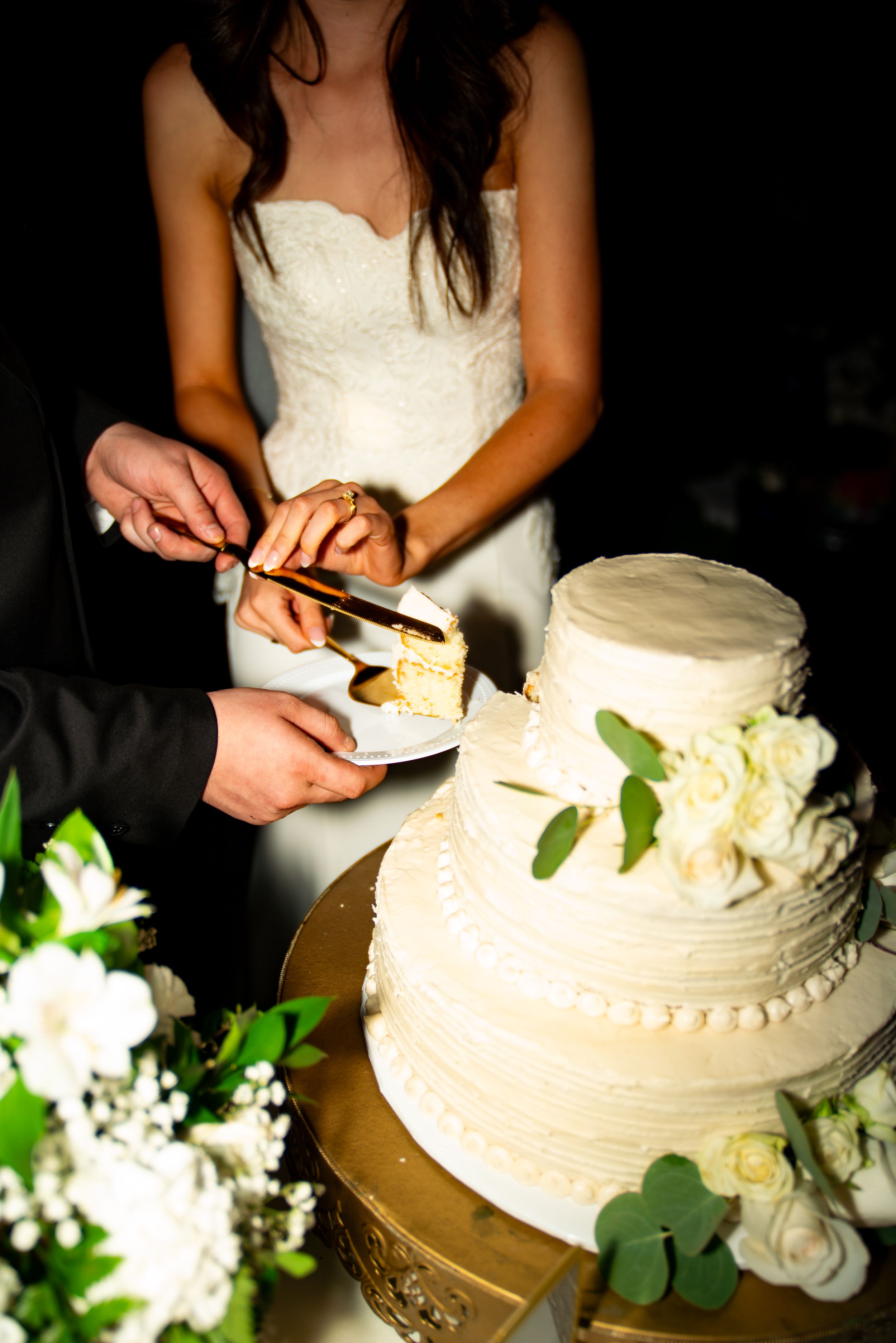White wedding cake being cut with decorative white flowers and greenery on display.