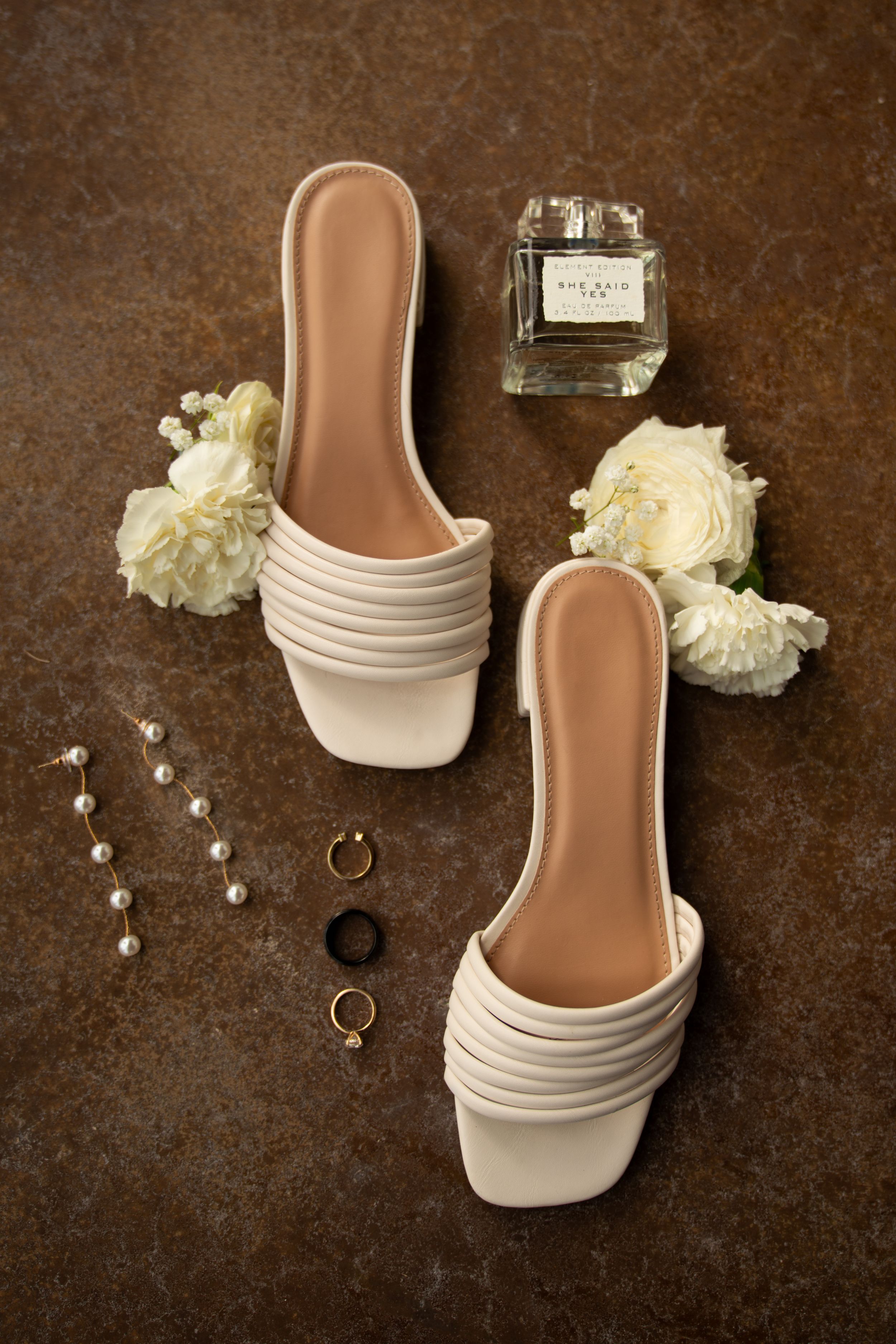 White bridal shoes and wedding rings displayed with floral accents on brown surface.