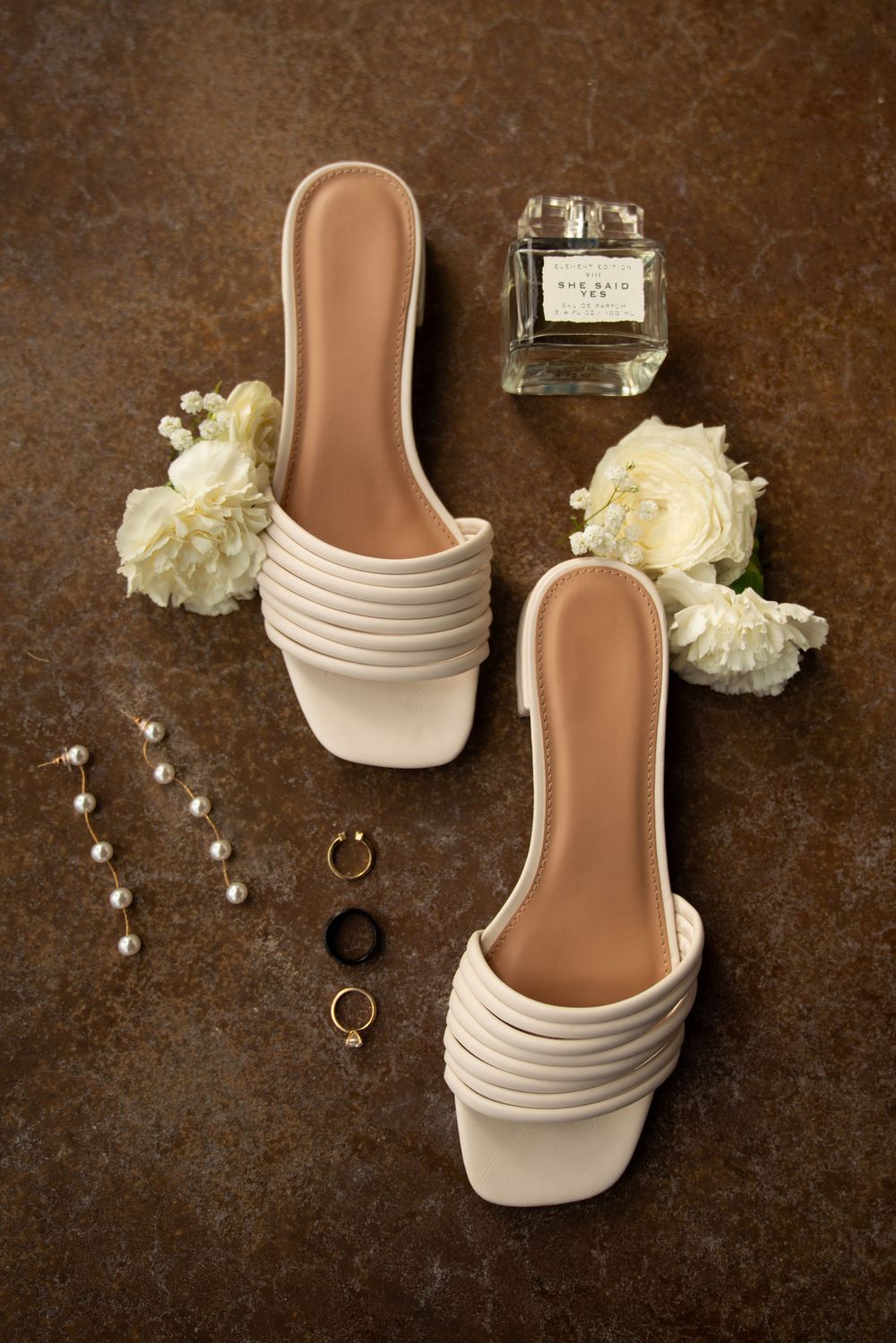White bridal shoes and wedding rings displayed with floral accents on brown surface.