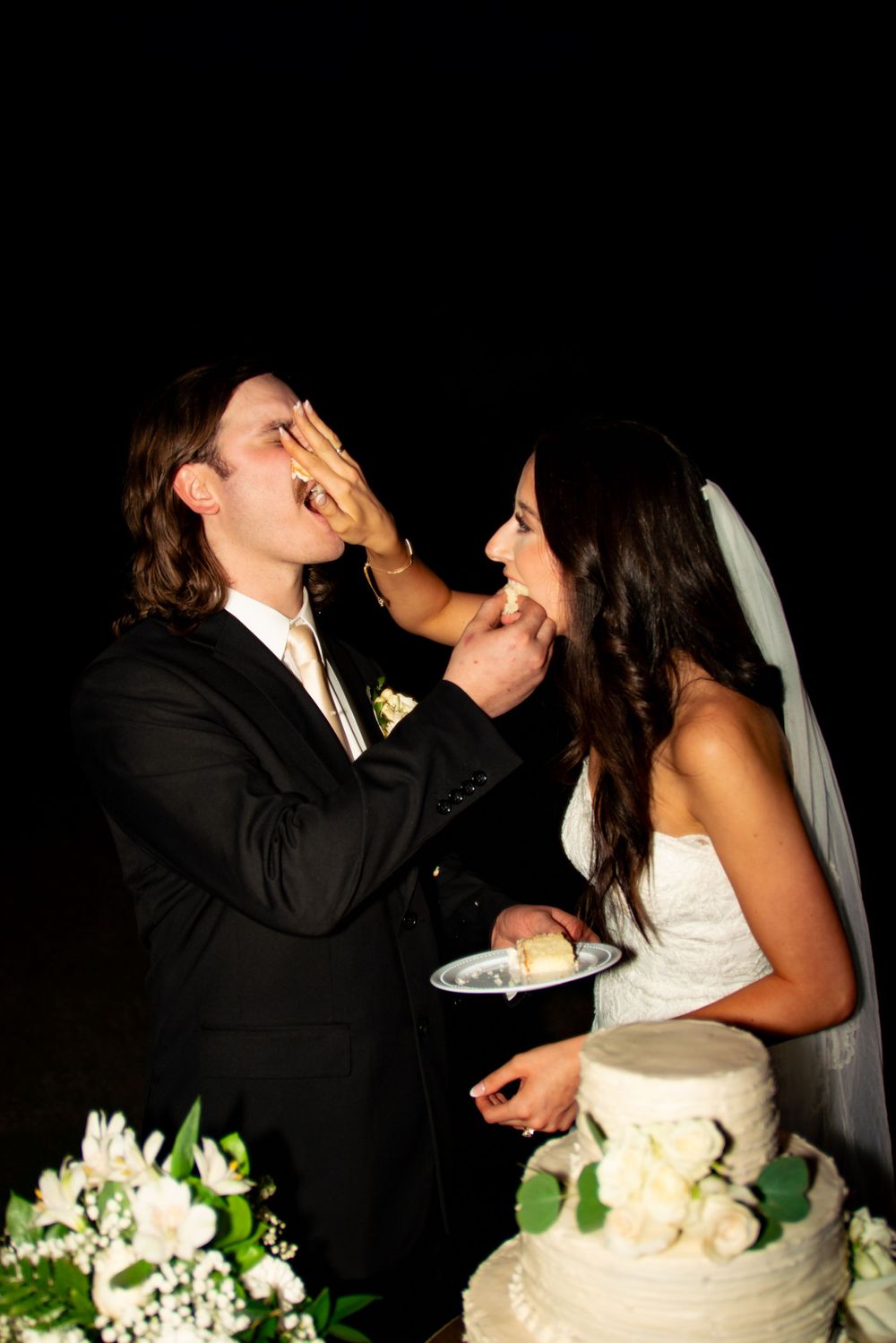 Newlyweds playfully feeding each other wedding cake at their reception.