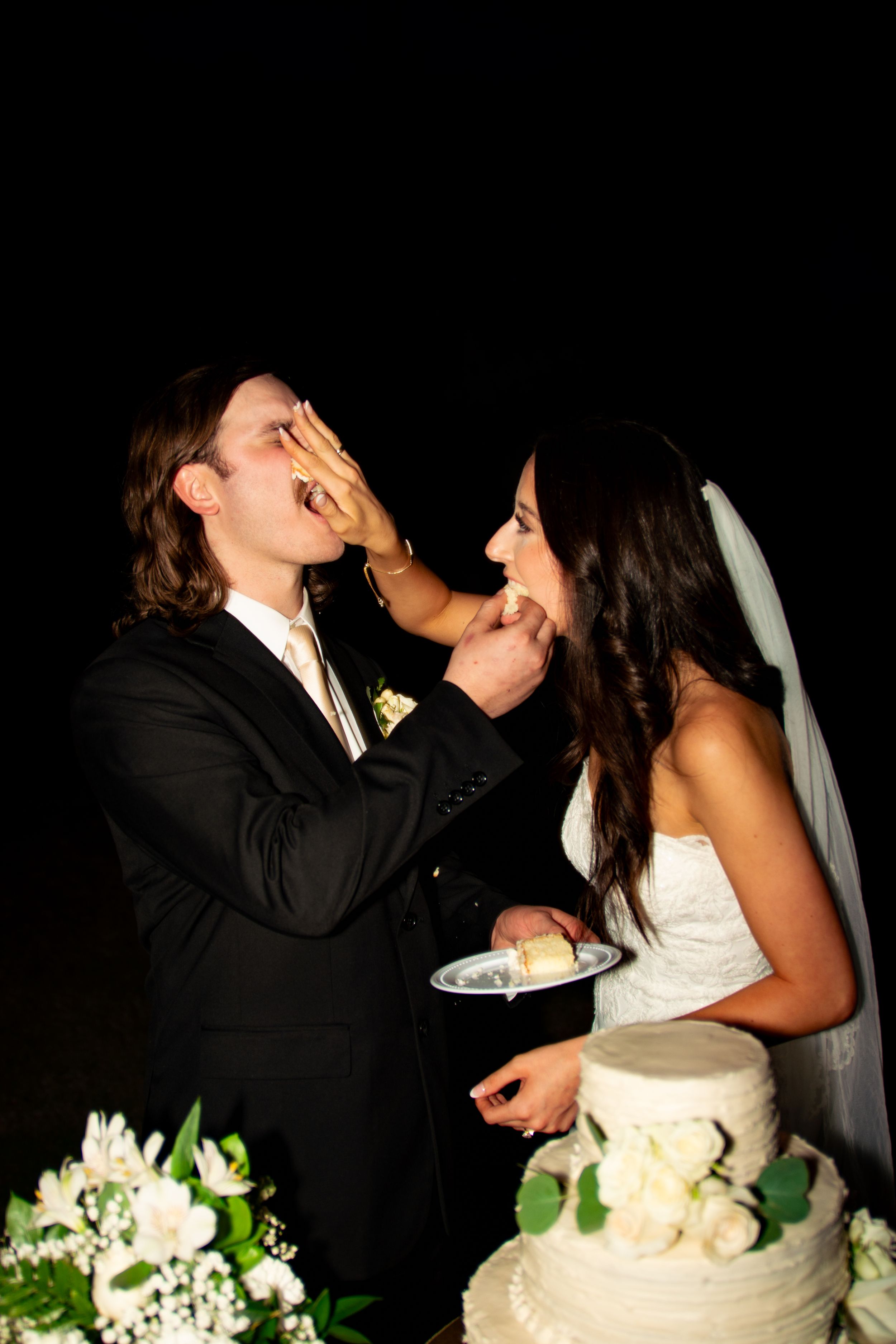 Newlyweds playfully feeding each other wedding cake at their reception.