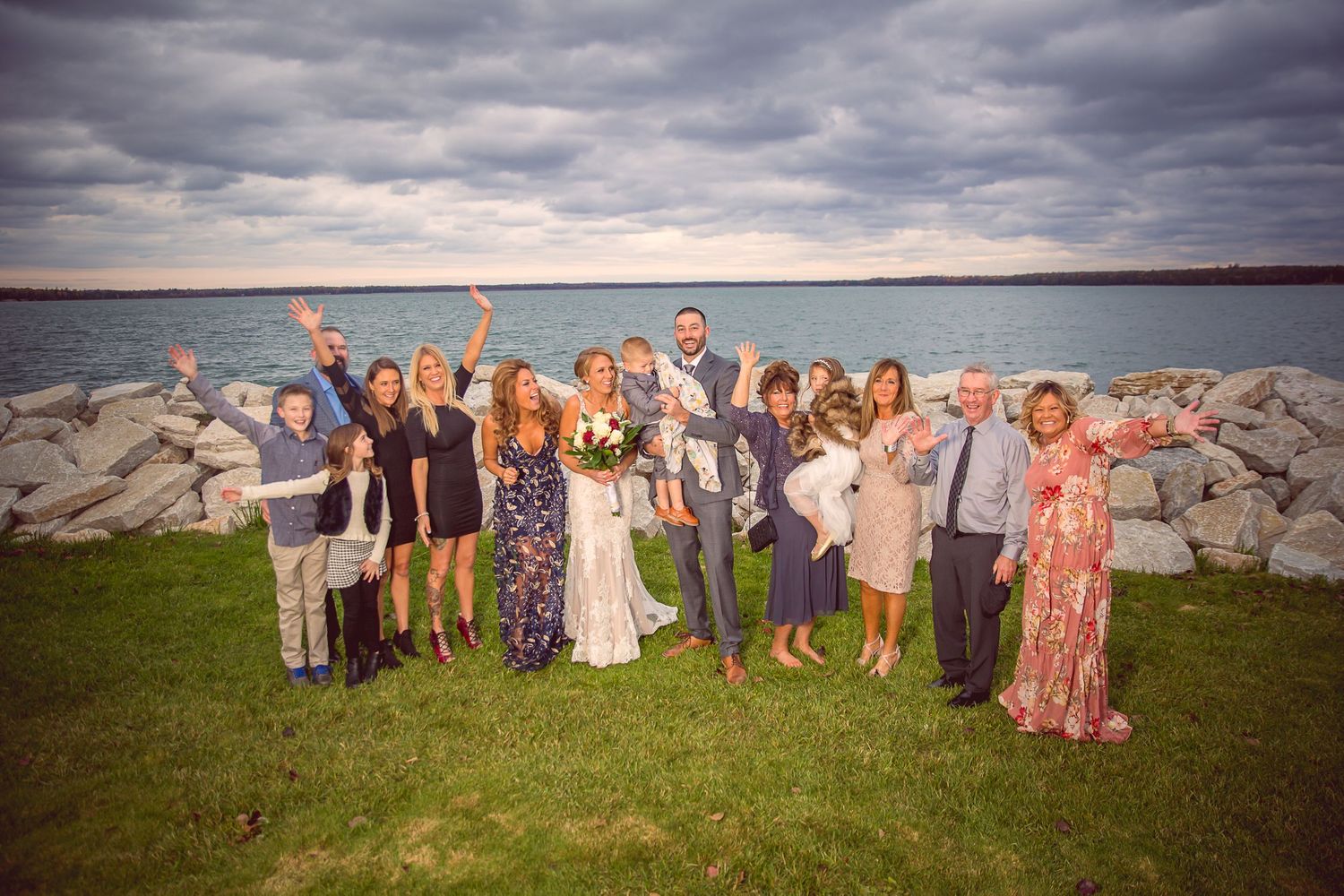 A joyful wedding group photo by the waterfront with dramatic cloudy skies.