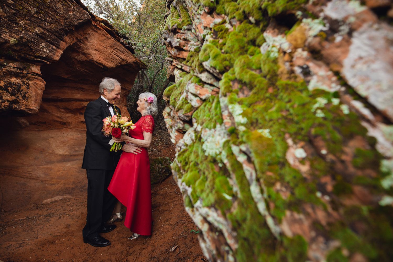 Senior couple in wedding attire share a tender moment in a mossy red rock canyon passage.