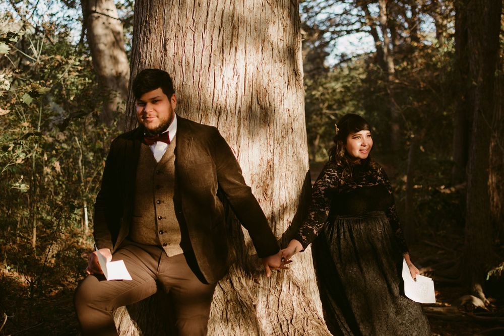 Couple in Victorian-inspired clothing hold hands while posing back-to-back against a massive tree in moody forest setting.