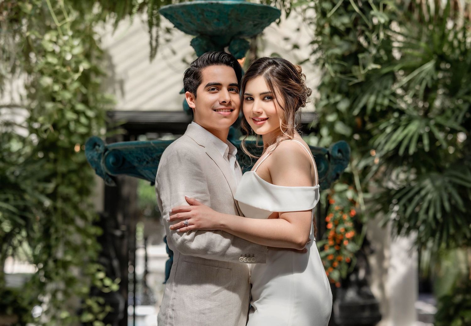 Romantic couple posing together outdoors near a fountain, dressed in wedding attire among lush greenery.