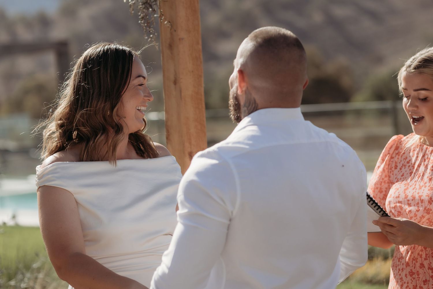 Wedding ceremony with wooden arch and floral arrangements against a natural landscape.